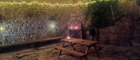 A stone-walled outdoor area with a wooden table and benches, a chimney, and string lights.