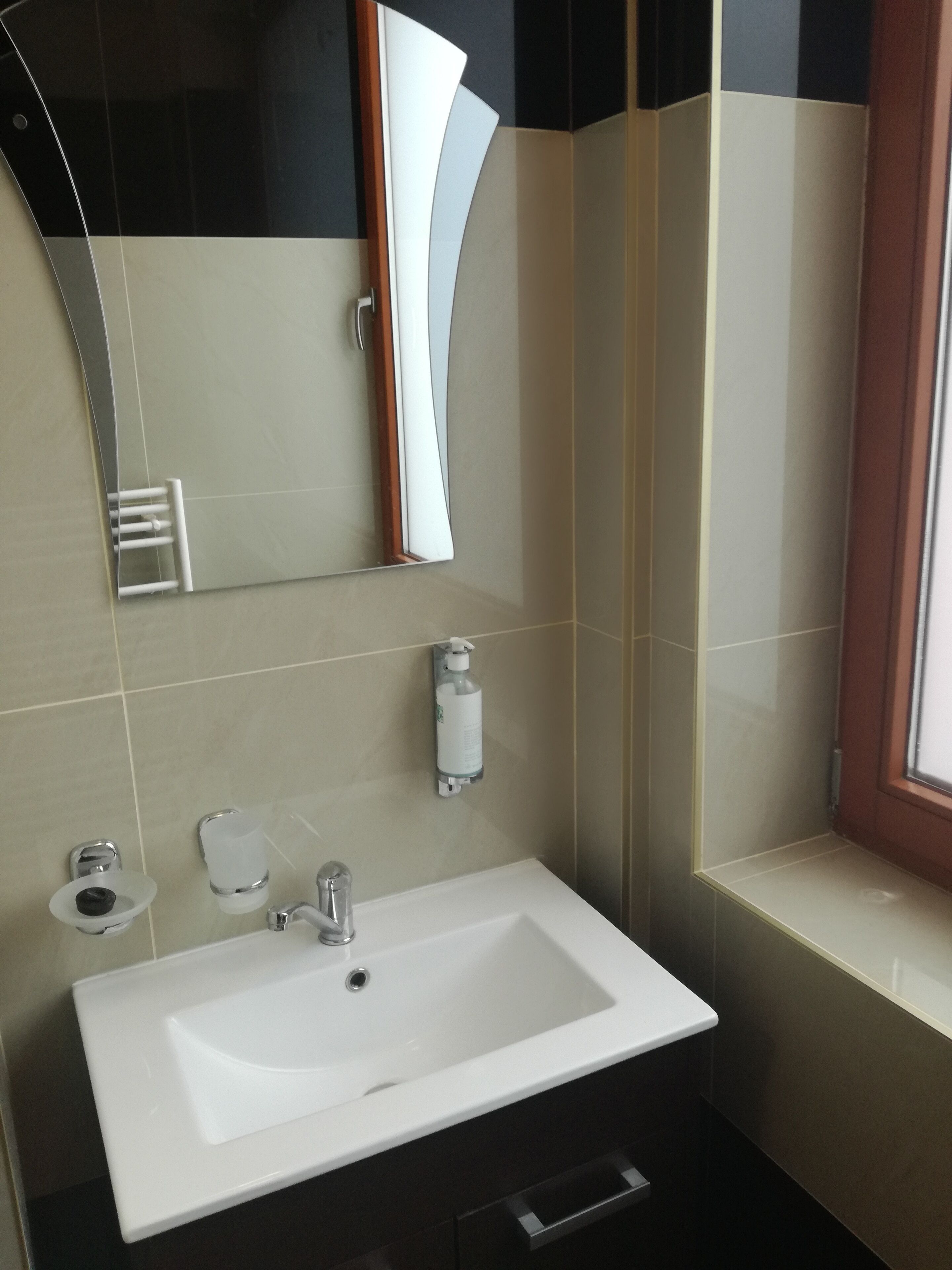 Une salle de bain avec un lavabo blanc, un miroir, un distributeur de savon et une fenêtre.