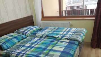Een bed met een geruit beddegoed en kussens, een raam met uitzicht op een balkon, en een radiator onder het raam.