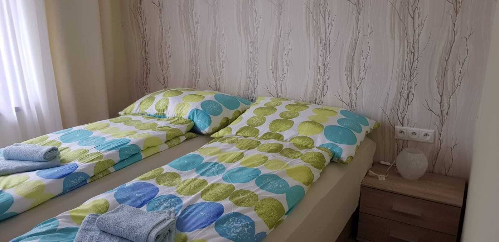 Una cama doble con colcha de lunares en verde, azul y amarillo. Dos almohadas con un diseño similar. Una toalla doblada está sobre la cama.