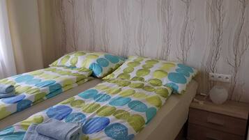 Una cama doble con colcha de lunares en verde, azul y amarillo. Dos almohadas con un diseño similar. Una toalla doblada está sobre la cama.