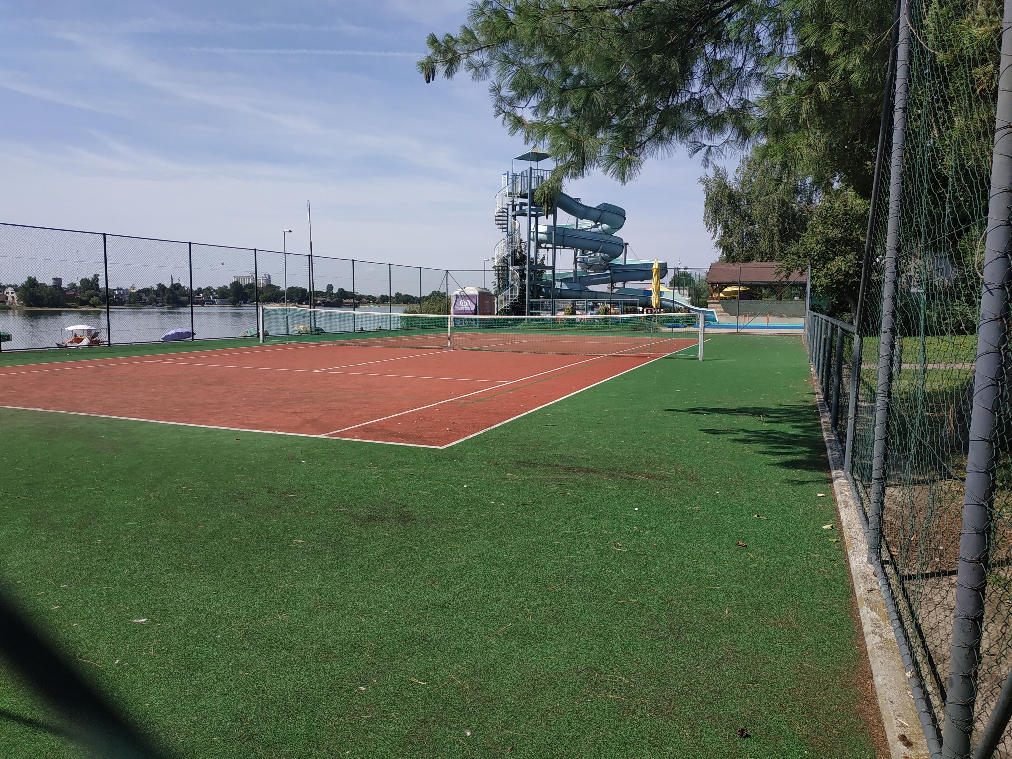 A tennis court with a net, surrounded by a fence, and a water slide in the background.
