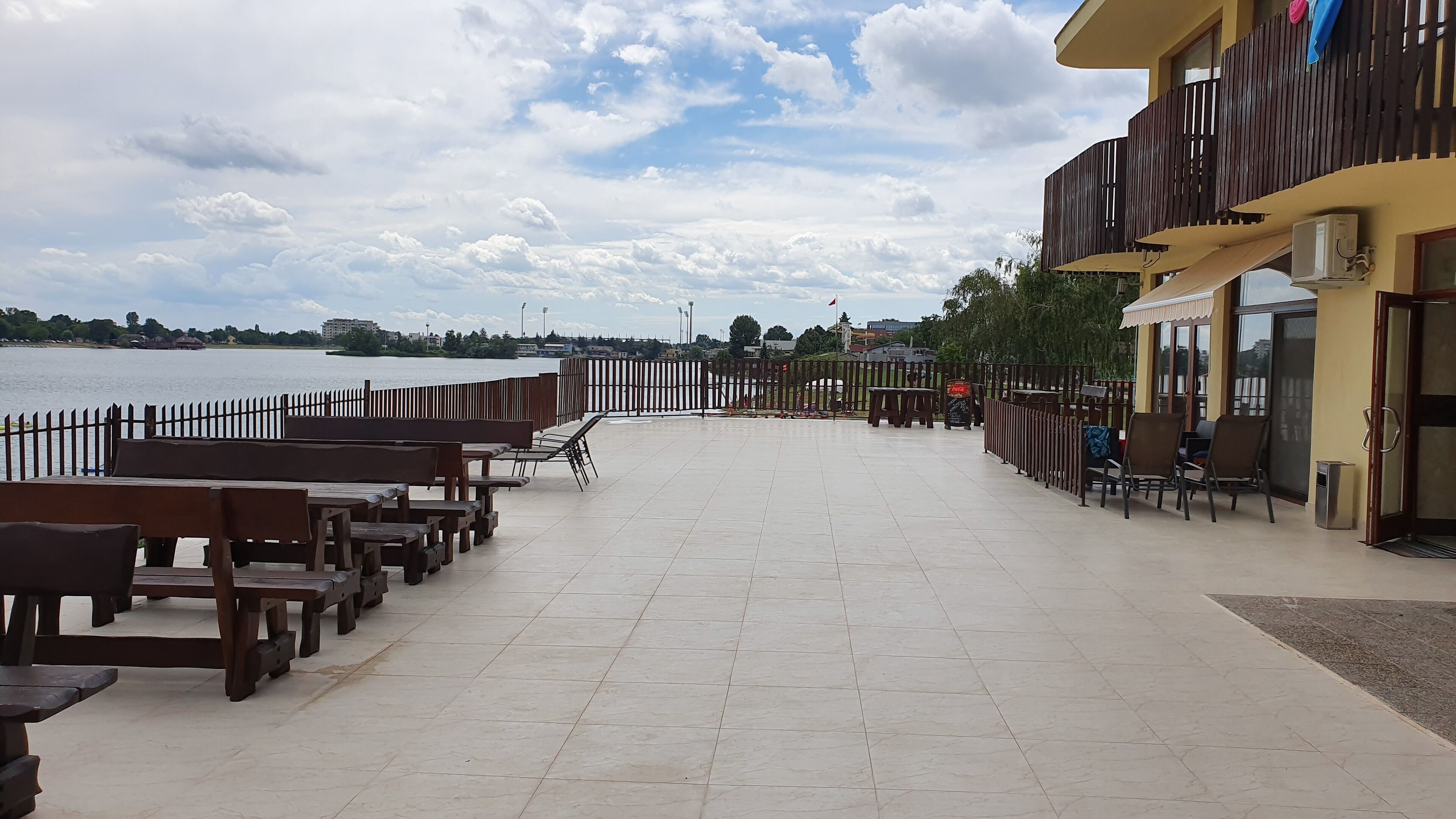 Terraza frente al agua con bancos y mesas de madera, piso de baldosas y vista al mar.
