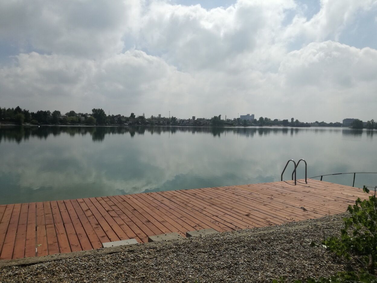 Un muelle de madera que se adentra en un lago tranquilo con un horizonte urbano al fondo.
