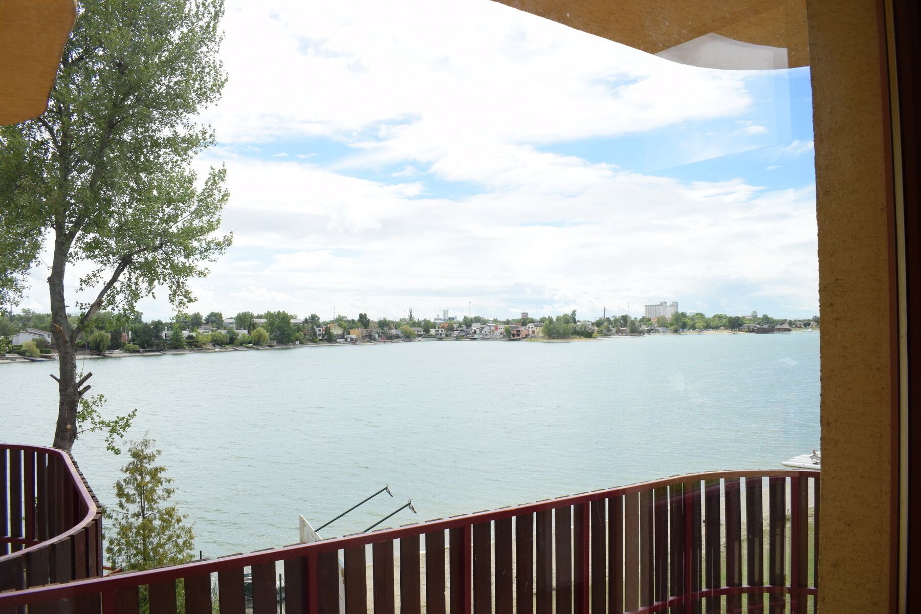 A view from a balcony overlooking a calm river with a distant shoreline, a tree in the foreground, and a clear sky.