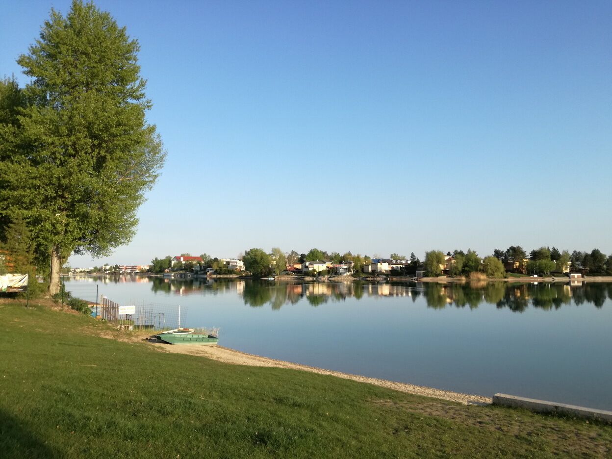 Un lago tranquillo con una riva erbosa, un molo e case sullo sfondo.
