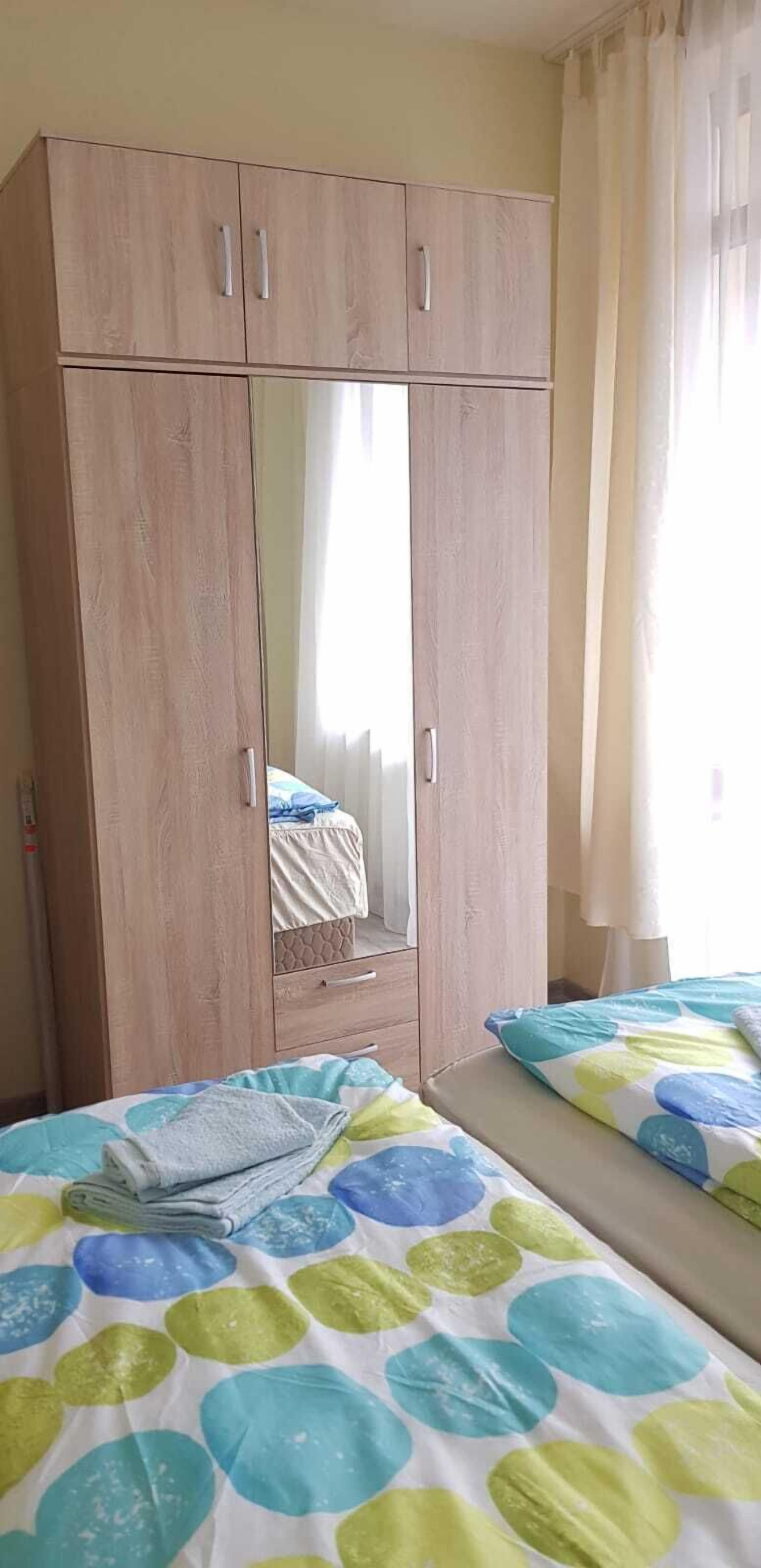A bedroom with a wooden wardrobe, a bed with a colorful bedspread, and a window with curtains.
