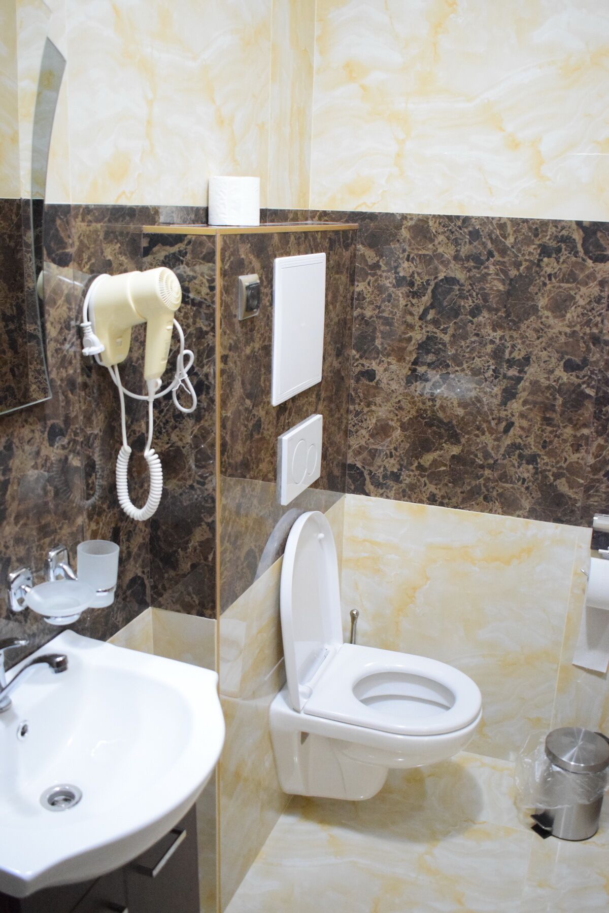 A bathroom with a marble wall, a white sink, a toilet, and a hairdryer mounted on the wall.