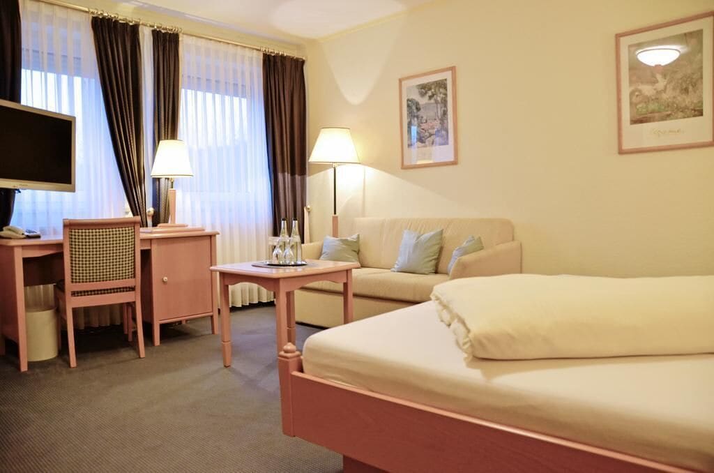 A hotel room with a bed, a desk with a chair, a television, a lamp, and a window with curtains.