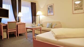 A hotel room with a bed, a desk with a chair, a television, a lamp, and a window with curtains.