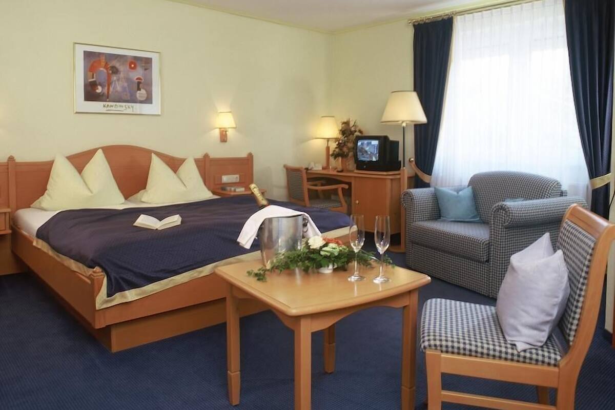 A hotel room with a bed, a desk with a computer, a chair, a table with a vase and glasses, and a window with curtains.