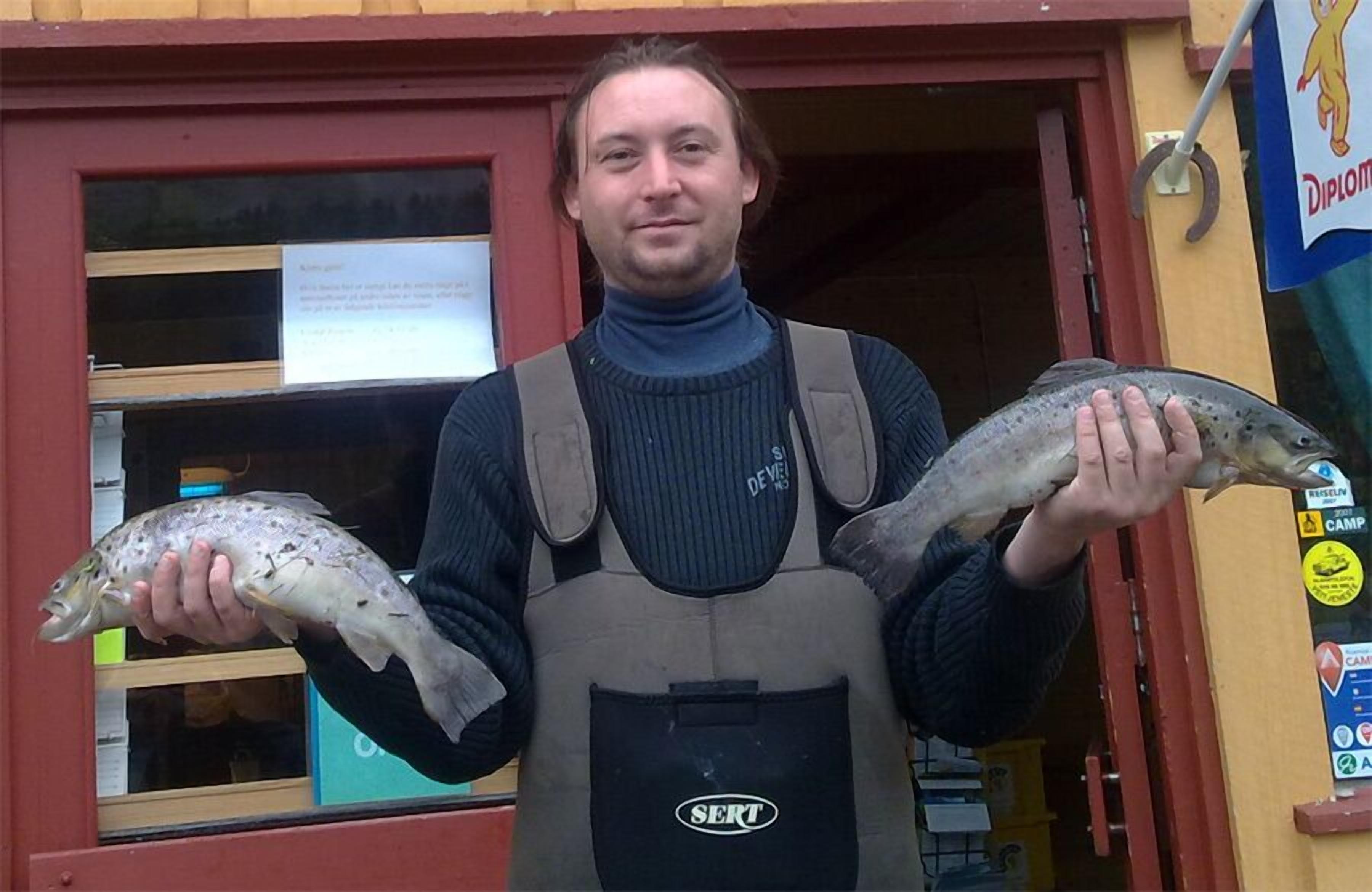 A person holding two fish in front of a building with a red door and a sign that reads "Diplom".