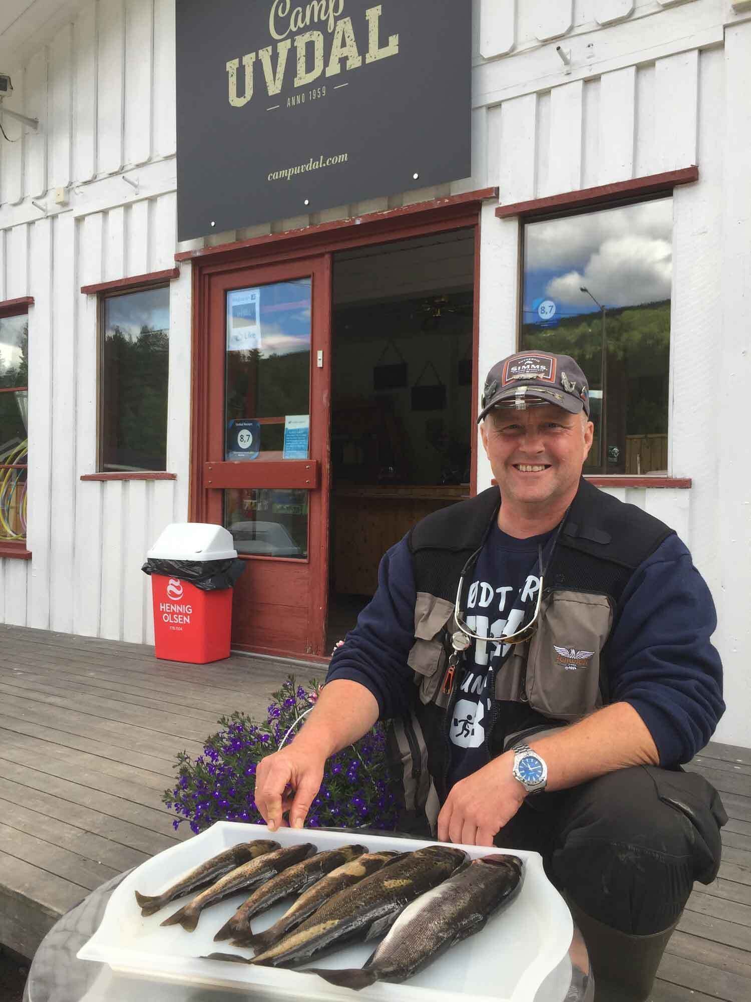 A man holding fish in front of a building with a sign that reads "Camp UVDAL".