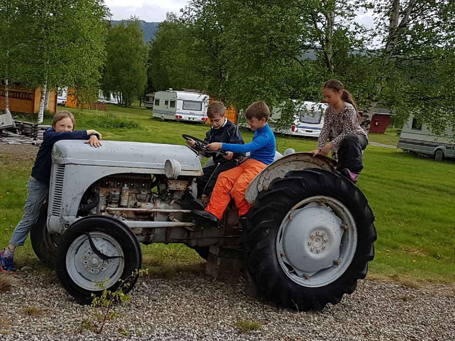 Ein alter Traktor mit vier Personen darauf, geparkt auf einem Kiesweg. Im Hintergrund Gras und Bäume.