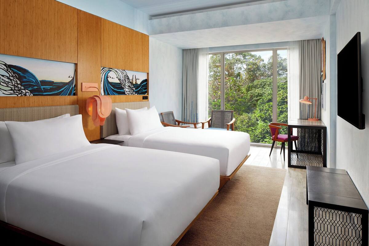 A hotel room with two beds, a desk, a chair, a TV, and large windows with a view of greenery.