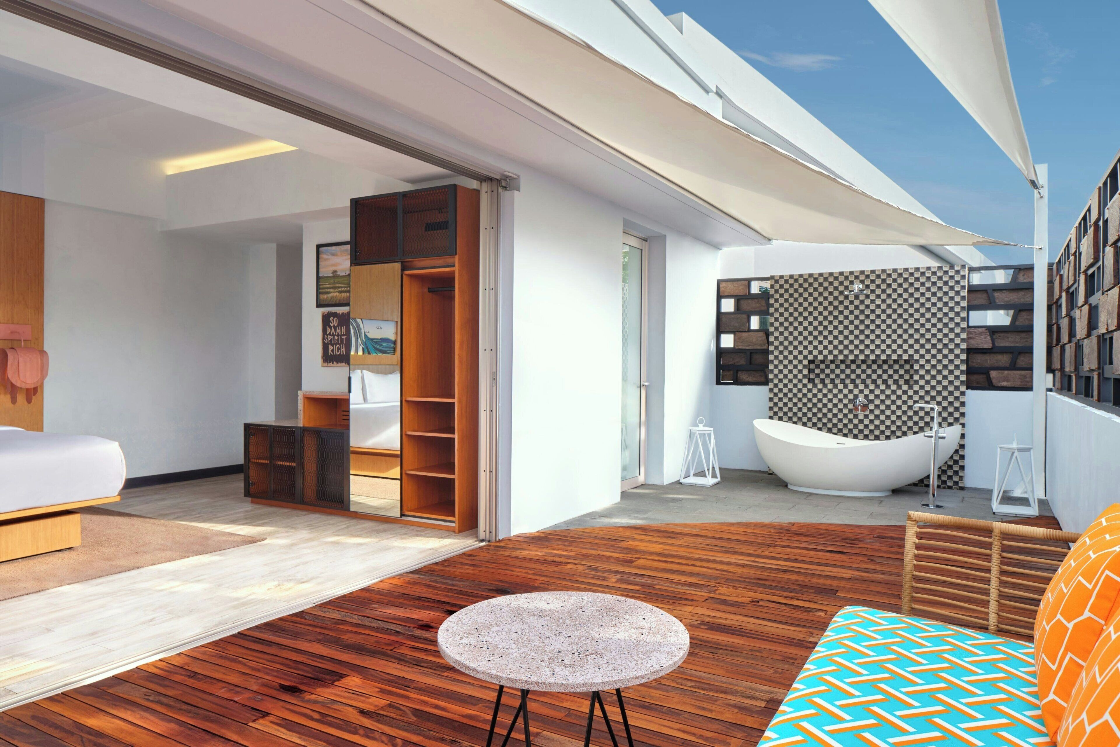 A modern interior with a wooden floor, a round stone table, a sofa with patterned cushions, a large white bathtub, and a view of the sky.