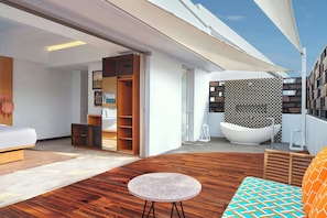 A modern interior with a wooden floor, a round stone table, a sofa with patterned cushions, a large white bathtub, and a view of the sky. - Aloft by Marriott Bali Seminyak (Keroboken Kelod)