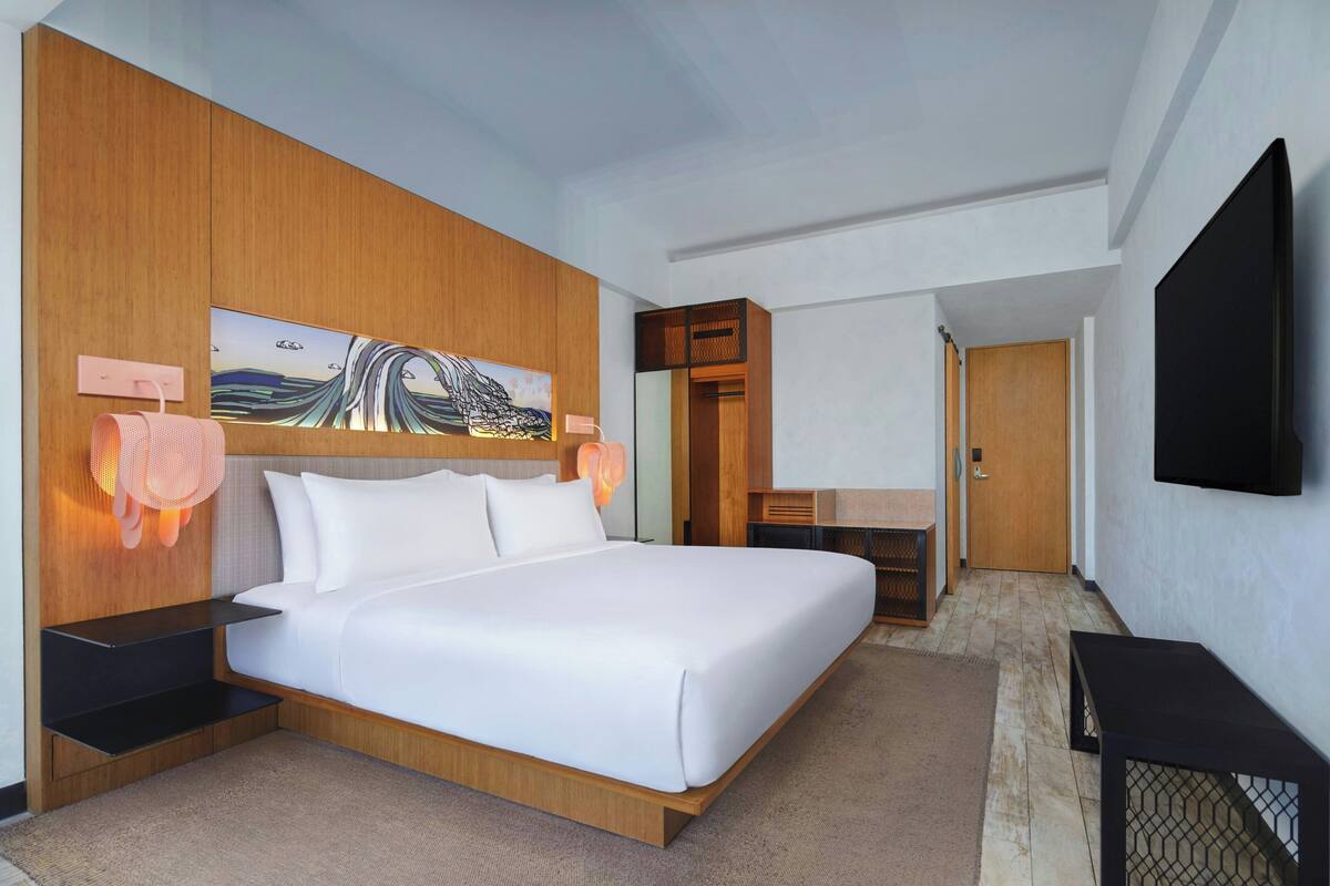 A modern hotel room with a large bed, wooden headboard, artwork, and a flat-screen TV.