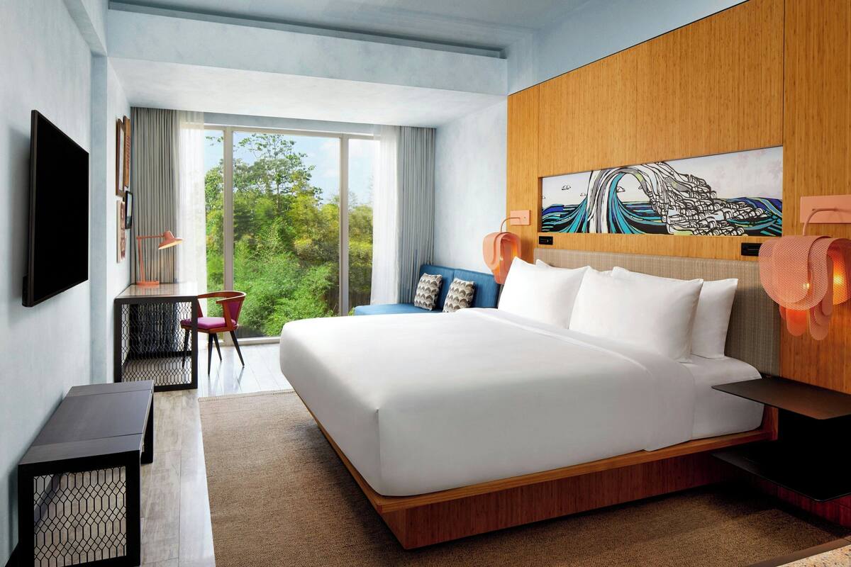 A modern hotel room with a large bed, a desk, and a chair. There is a large window with a view of greenery outside.
