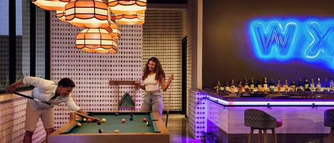 A pool table with two players, a bar area with a neon sign, and patterned wall decor.
