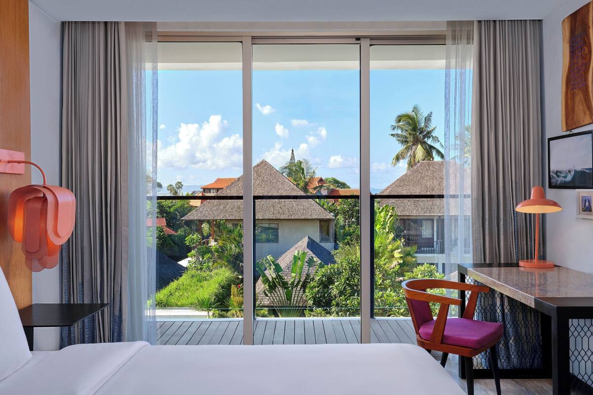 A hotel room with a large sliding glass door leading to a balcony, a bed, a desk with a chair, and a lamp.