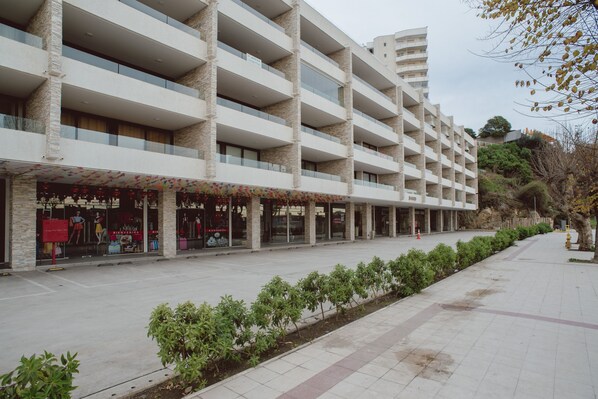 A multi-story hotel with a modern design, featuring balconies on each floor, a covered walkway, and a landscaped area with small trees.