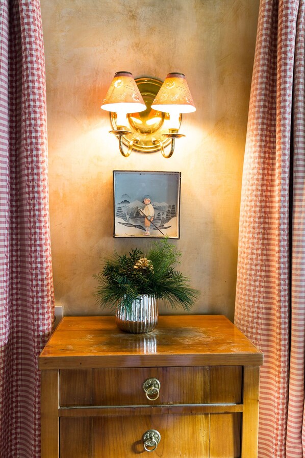 A wooden nightstand with a vase of greenery, a framed picture, and a wall-mounted lamp. - Hotel Edelweiss (Lech am Arlberg)