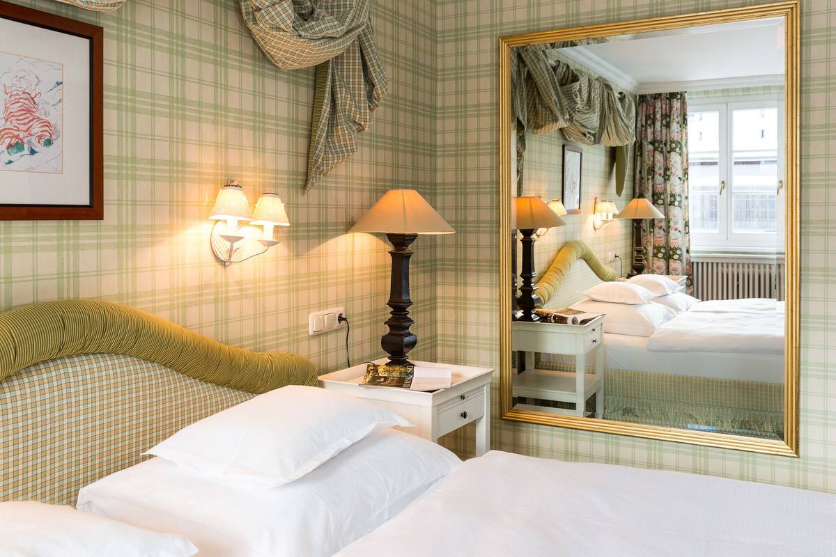 A hotel room with a checkered wallpaper, two beds with white linens, a bedside table with a lamp, a mirror, and a window with curtains.