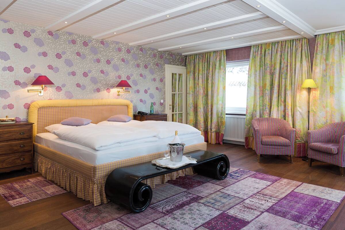 A bedroom with a large bed, two bedside lamps, a nightstand, a wooden dresser, a patterned rug, and a window with curtains.