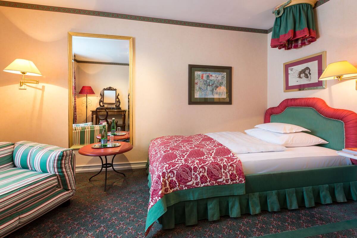 A hotel room with a bed, a sofa, a table with a lamp, and a mirror.
