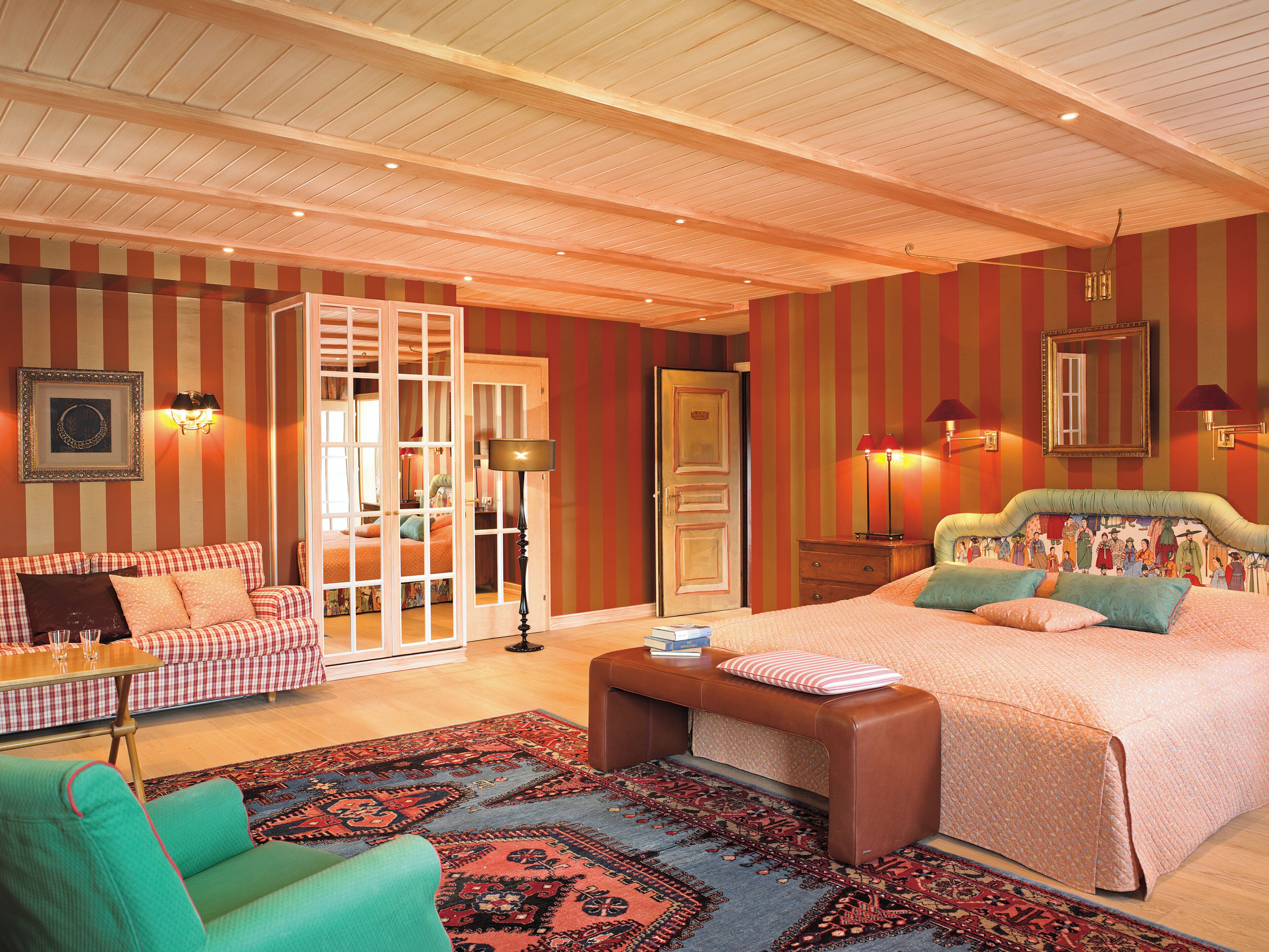 A bedroom with a large bed, a sofa, a chair, and a desk. There is a door leading to another room.