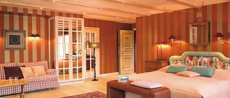 A bedroom with a large bed, a sofa, a chair, and a desk. There is a door leading to another room.