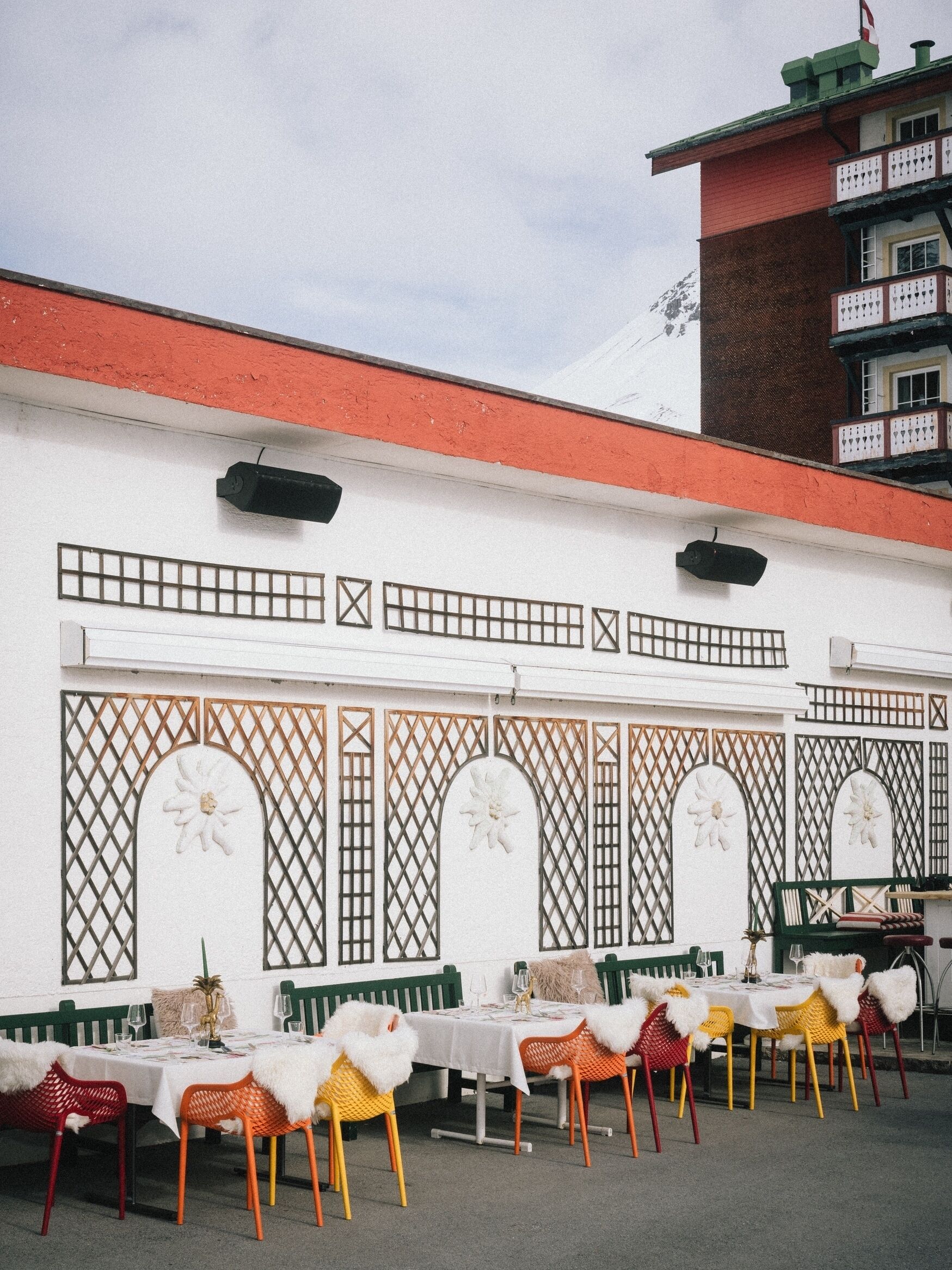 A dining area with outdoor tables and chairs, a white wall with decorative panels, and a building with a red roof in the background.
