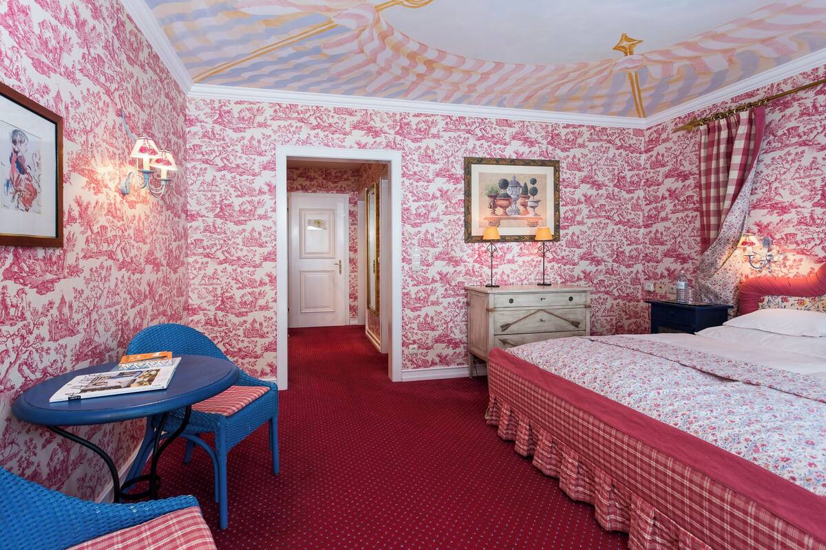 A room with floral wallpaper, a bed with a red and white checkered bedspread, a blue chair with a magazine, and a nightstand with a lamp.