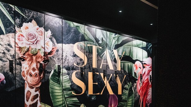 Una habitación con un mural grande que muestra una jirafa y flamencos, un banco y un letrero que dice 'STAY SEXY'.