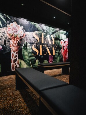 A room with a large wall mural featuring a giraffe and flamingos, a bench, and a sign that reads "STAY SEXY". - Hotel Edelweiss (Lech am Arlberg)