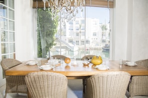 A dining area with a wooden table set for four, surrounded by wicker chairs, with a view of buildings through large windows. - Villa Marina (Hammamet)