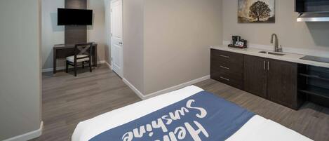 A hotel room with a bed, a desk, a chair, a TV, a painting, and a kitchenette.