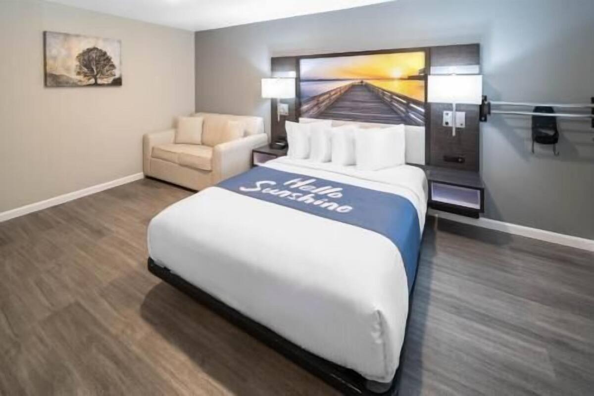 A hotel room with a bed, a chair, a nightstand, a lamp, and a wall art piece.