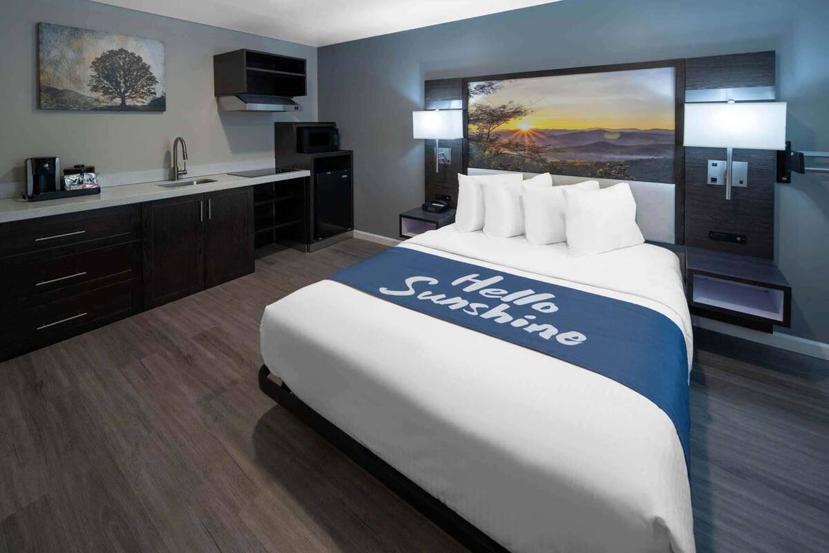 A modern hotel room with a bed, a nightstand, a desk with a computer, a microwave, and a painting on the wall.