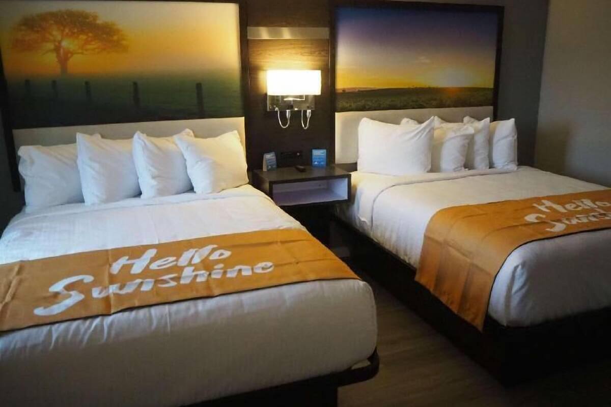 A hotel room with two beds, each with a 'Hello Sunshine' bedspread, a nightstand with a lamp, and a wall-mounted light.