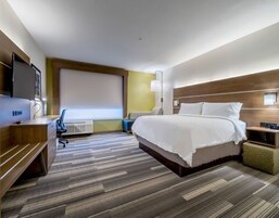 A modern hotel room with a large bed, a desk, a chair, a TV, and a window with blinds.