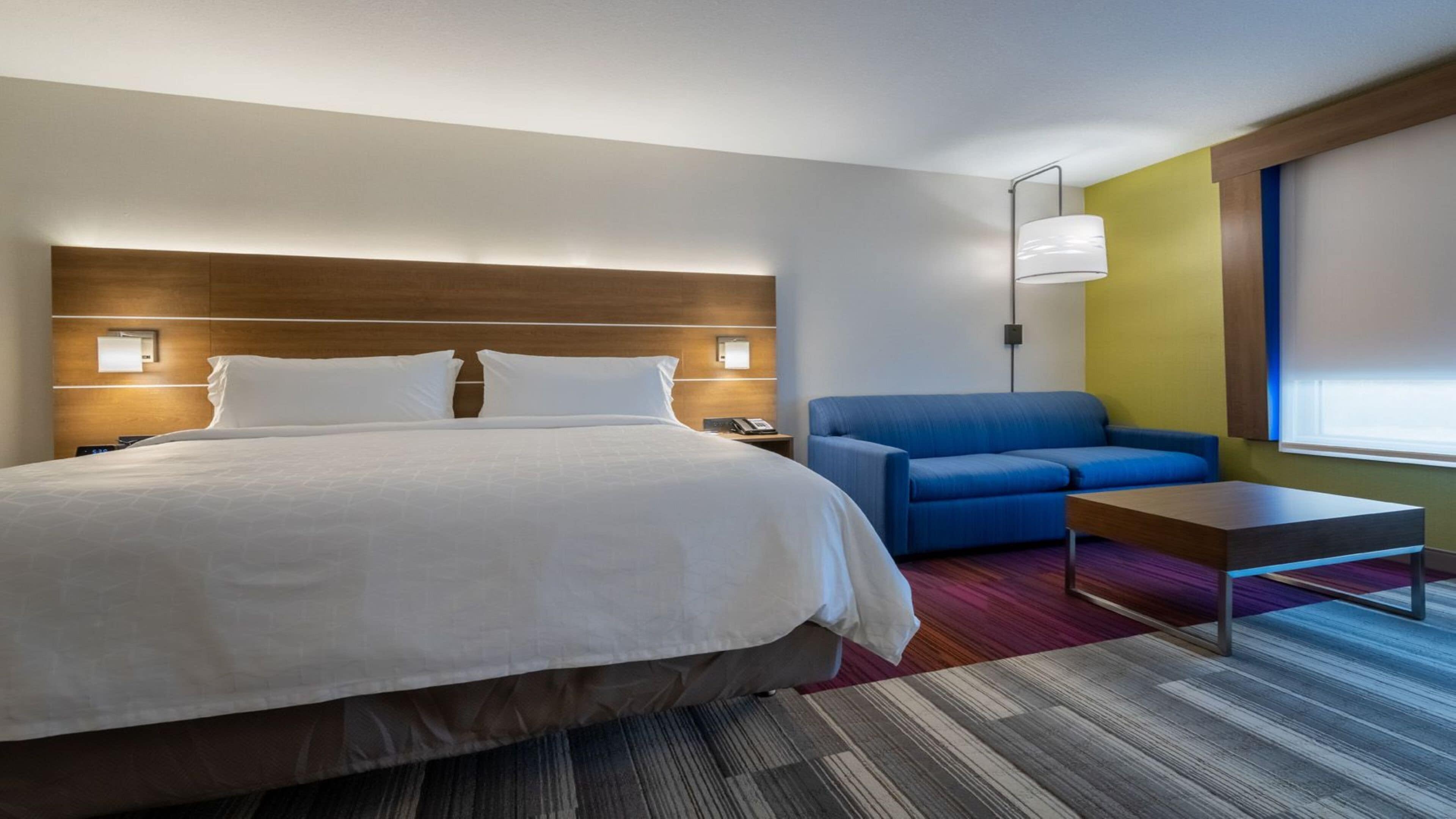 Una habitación de hotel con una cama grande, un sofá azul, una mesita pequeña y una mesita de noche con una lámpara.