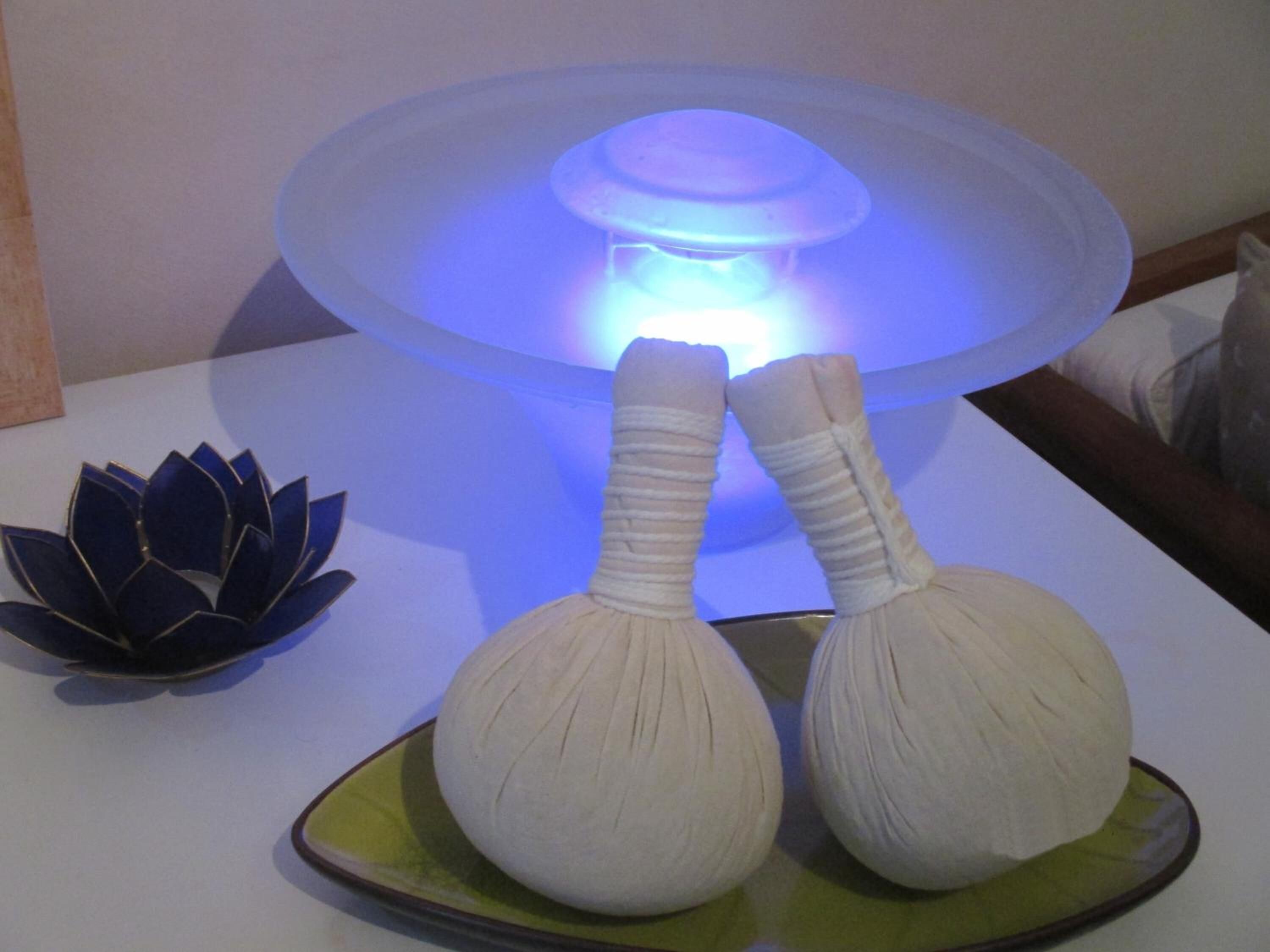 Una mesa con una lámpara azulada, dos objetos blancos que parecen herramientas de masaje, y una flor de loto decorativa.