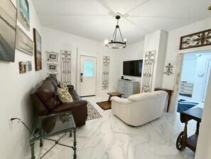 Un salón con un sofá de cuero marrón, un sofá blanco, una mesa de centro de vidrio, un televisor y decoración en la pared.