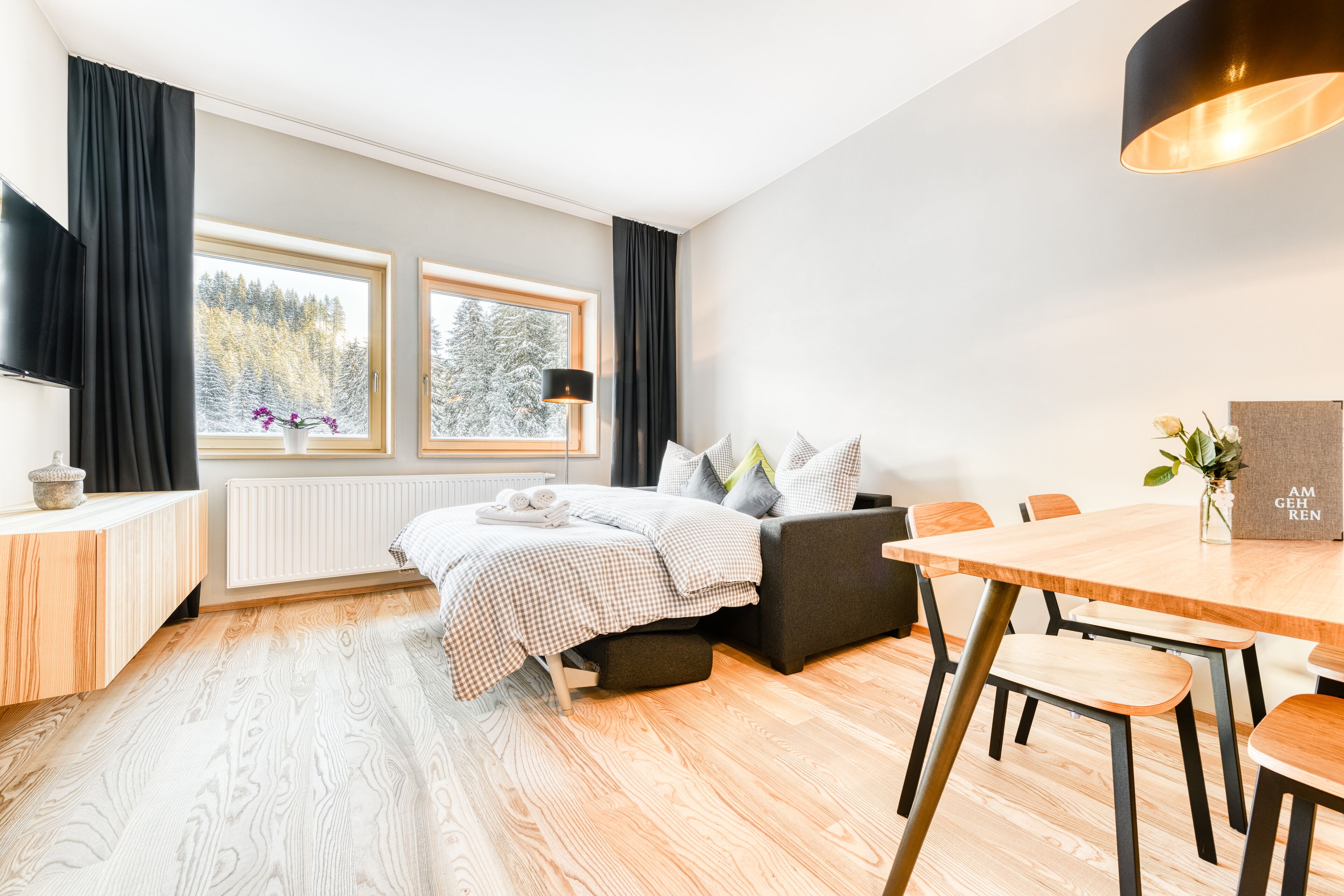 Ein modernes Hotelzimmer mit einem Bett, einem Esstisch und einer Couch. Zwei Fenster mit Vorhängen, ein Heizkörper und ein Fernseher befinden sich auf der linken Seite.