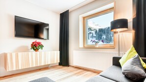 A modern living room with a grey sofa, a mounted TV, a window with a view of snow-capped mountains, and a floor lamp.