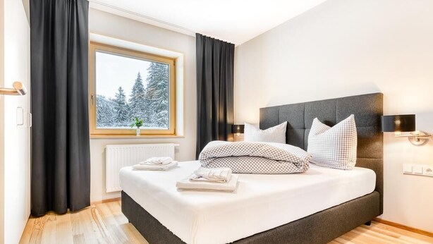 A modern bedroom with a large bed, black curtains, a window with a view of snow-covered trees, and a radiator.