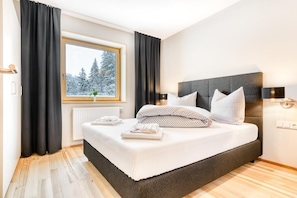 A modern bedroom with a large bed, black curtains, a window with a view of snow-covered trees, and a radiator.