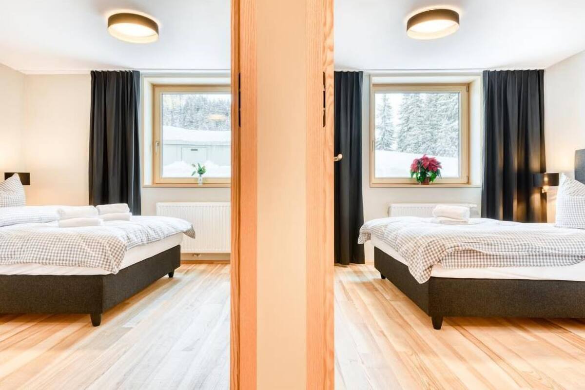 A mirrored view of a bedroom with two beds, large windows with curtains, a radiator, and a potted plant.
